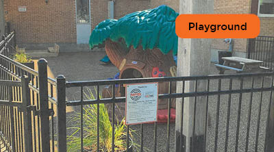 A gated outdoor recycled rubber play area with play structures outside of a brick building.
